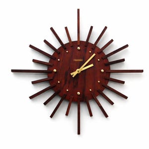 May include: A mid-century modern wall clock with a sunburst design. The clock features a dark brown wooden face with gold-colored hands and hour markers. The clock's rays are made of dark brown wood and extend outward from the center.