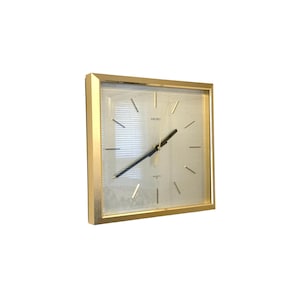 May include: Square gold-framed Seiko quartz clock. The clock face is a light color with black hands and markers. The clock has a minimalist design, with the word "SEIKO" printed on the face.