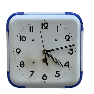 May include: Square white alarm clock with a blue frame. The clock face has black numbers and hands, with a small gold-coloured knob at the centre. The clock is set to approximately 3:20.