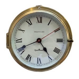 Puede incluir: Un reloj redondo de color latón con esfera blanca y números romanos negros. Las manecillas son negras y rojas. El texto "WESTCLOX" y "Quartzmatic" son visibles en la esfera. Reloj de estilo vintage.
