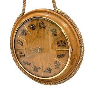 May include: A round wall clock with a light wood grain face featuring twelve metallic zodiac symbols as hour markers. The clock has a gold-toned metal rim and a braided tan cord border. The centre displays thin gold hands and a small logo.