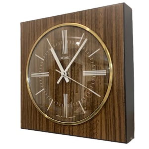 Puede incluir: Un reloj de pared cuadrado con acabado de veta de madera, que presenta un bisel circular dorado alrededor de una esfera marrón. El reloj muestra marcadores de hora blancos, manecillas blancas y el texto Metamec y Made in England en la esfera.