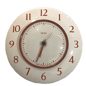 Puede incluir: Un reloj de pared redondo con esfera blanca y un borde ancho de color blanco. El reloj presenta números rojos del 1 al 12, marcas de minutos rojas y manecillas rojas. La palabra SMITHS está impresa en rojo y MADE IN GREAT BRITAIN en la base.