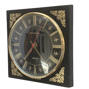 May include: A square black wall clock with a gold-colored circular face. The clock face features gold Roman numerals, gold hands, and the word "STANDARD". Decorative gold filigree accents the corners. The clock is labeled "REPUBLIC OF IRELAND".