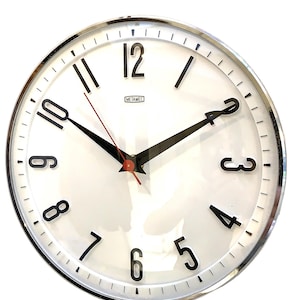 May include: A round wall clock with a polished chrome frame and a white face. The dial features black Arabic numerals for 3, 4, 5, 6, 8, 10, and 12, with black hour and minute hands, a thin red second hand, and the brand name Metamec printed in black.