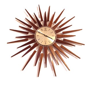 Peut inclure: Une horloge murale en forme d'éclat d'étoile de style moderne du milieu du siècle, avec un cadran argenté et des rayons en bois marron. L'horloge a un cadran circulaire avec des chiffres et des aiguilles noirs.