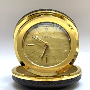 May include: A round Europa travel alarm clock with a gold-tone brushed metal face and casing. The clock features gold hands, hour markers, and the text Europa and Germany printed on the dial. It is housed in a dark brown textured circular case.