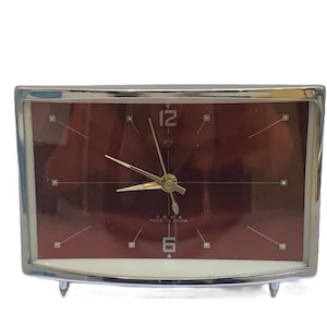 May include: A vintage rectangular clock with a maroon face and silver-colored frame. The clock face features gold-colored hands and hour markers. The text "MADE IN CHINA" is visible at the bottom. The clock has two small feet.
