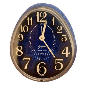 Può includere: Una sveglia musicale Goldbühl a forma di uovo con quadrante blu glitterato, numeri bianchi e sottili lancette dorate. L'orologio presenta un bordo blu testurizzato e le scritte Goldbühl, MUSICAL ALARM e WEST GERMANY sul quadrante.