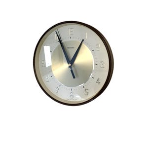 May include: A circular wall clock with a silver face and a dark brown frame. The clock features black hands and a metallic silver centre. The numbers are in a clear, easy-to-read font. The clock face has the text "METAMEC" and "MADE IN ENGLAND".