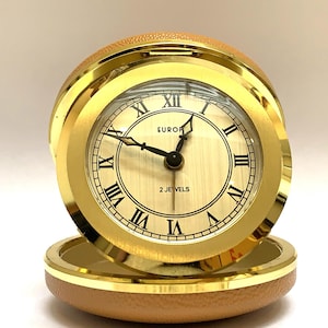 May include: A round travel clock with a gold-tone metal frame and a tan textured leather-like case. The clock face features a cream-coloured background with black Roman numerals, black hands, and the text EUROPA and 2 JEWELS printed on the dial.