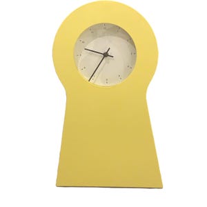 May include: A yellow table clock with a keyhole-shaped frame and a circular white face. The clock features black hour, minute, and second hands, with black numerical markings from one to twelve positioned around the perimeter of the white dial.