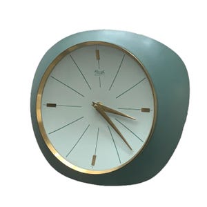 May include: A vintage wall clock with a teal-colored, asymmetrical frame. The clock face is white with gold-colored hands and a gold-colored rim. The clock has the text "Kieninger" and "WATERMATIC" on the face.