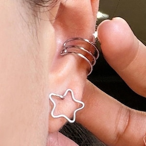 May include: A silver ear cuff with three connected bands and a star-shaped earring. The jewelry is worn on the ear. The star earring is a simple outline of a five-pointed star.