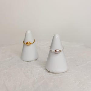 May include: Two rings displayed on white ceramic cones. One ring features a gold band with a round, golden-colored pearl. The other ring has a silver band with a light pink, oval-shaped pearl. Both rings are set against a white background.