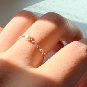 May include: A delicate ring featuring a thin silver-toned metal chain band with a single small, round, white pearl centered on the front. The chain links are interconnected, creating a flexible and lightweight band that encircles the finger.