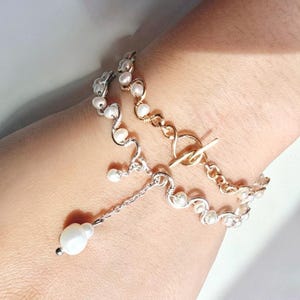 May include: A delicate bracelet featuring freshwater pearls and a toggle clasp. The bracelet has a silver and gold tone design with a dangling pearl charm. The jewelry is designed for a feminine aesthetic.