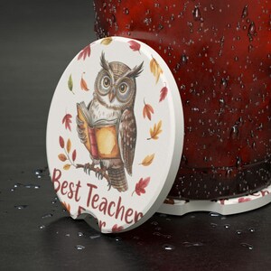 May include: White car coaster with an owl illustration reading a book, surrounded by autumn leaves. The text "Best Teacher Ever" is printed in a script font. The coaster is designed to fit in a car cup holder.