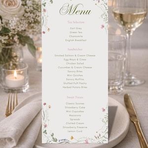 Può includere: Un menu di nozze con un design floreale, che elenca selezioni di tè, panini e dolci. Il menu è su sfondo bianco, con un bicchiere di vino e una candela. Il menu include tè Earl Grey, mini cupcake e torta alle fragole.
