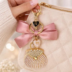May include: A gold-toned keychain featuring a dusty pink satin bow, a black heart charm with the letter R, a small white heart charm with a gold bow motif, and a dangling handbag charm encrusted with clear crystals and a central iridescent stone.