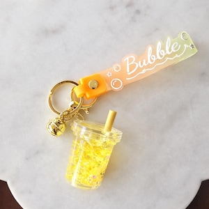 May include: A clear keychain featuring a miniature bubble tea cup filled with yellow glitter and a straw. The keychain has a gold-colored ring and a strap with the word "Bubble" in white lettering, with orange and yellow gradient.