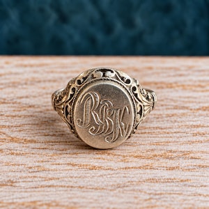 Op de afbeelding: Een zilverkleurige metalen ring met een ovaalvormig vlak voorzien van een gegraveerd monogram met de letters RBK. De ringband en de zetting rondom het centrale ovaal tonen gedetailleerd, opengewerkt filigraanwerk met een donkere patina.