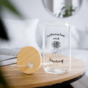Op de afbeelding: Een helder, blikvormig drinkglas met een bamboe deksel en rietje. Op het glas staan de woorden "Caffeinated and Creating" en een lijntekening van een persoon met een gekrabbelde kop. Het deksel en de tafel zijn van hout.