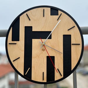 May include: A round wooden wall clock with a light wood face and black geometric design. The clock has black hands, a red second hand, and a black border. The clock is hanging on a metal railing.