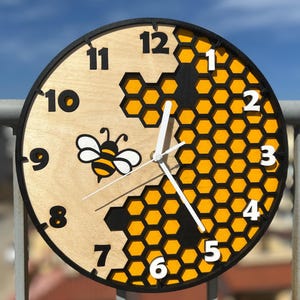 May include: A round wooden wall clock with a black frame and white hands. The clock face features a bee illustration and a honeycomb design in yellow and black. The numbers are black and the clock measures approximately 12 inches in diameter.