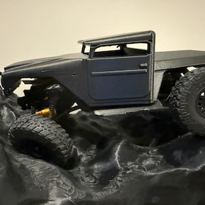 Puede incluir: Un modelo de camión todoterreno negro con neumáticos grandes y texturizados. El camión tiene una carrocería detallada, que incluye cabina, puertas y caja. El vehículo está posicionado sobre una superficie negra texturizada, lo que sugiere un terreno accidentado.