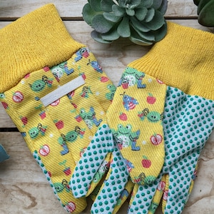 May include: A pair of yellow gardening gloves with a green and white polka dot pattern on the fingertips. The gloves have a cartoon print of green bugs and red apples. The gloves are labeled "Kids gloves Ages 3-6yrs".
