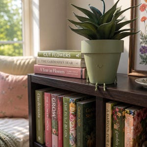 May include: A matte sage green ceramic plant pot with a simple embossed smiley face and two small dangling legs, containing a dark green succulent plant. The pot sits on a dark wooden shelf next to a stack of gardening books with various titles.