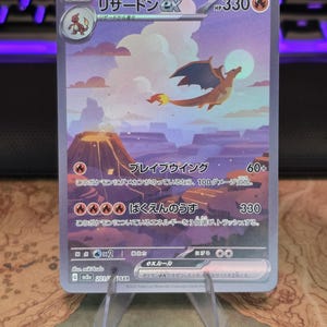 Pu&ograve; includere: Una carta collezionabile Charizard ex raffigurante una creatura alata che vola sopra un paesaggio vulcanico con luna piena. La carta mostra 330 HP, testo in giapponese e simboli di tipo fuoco su un cielo dai toni viola e arancioni.