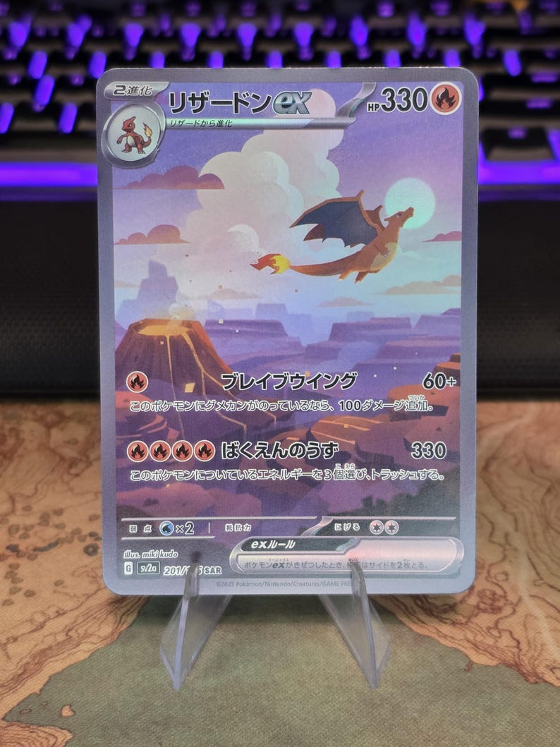 Pu&ograve; includere: Una carta collezionabile Charizard ex raffigurante una creatura alata che vola sopra un paesaggio vulcanico con luna piena. La carta mostra 330 HP, testo in giapponese e simboli di tipo fuoco su un cielo dai toni viola e arancioni.