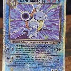 May include: A holographic Dark Blastoise trading card featuring the blue Shellfish Pokémon with two cannons on its back. The card displays 70 HP, the Hydrocannon and Rocket Tackle attacks, and lists the Pokémon's length as 5' 3" and weight as 189 lbs.