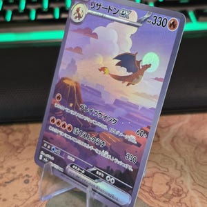 Pu&ograve; includere: Una carta collezionabile giapponese Charizard ex con un'illustrazione olografica del drago che vola sopra un paesaggio vulcanico notturno con la luna piena. La carta mostra 330 HP, simboli di energia fuoco e testo giapponese sulle mosse.