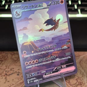 Pu&ograve; includere: Una carta collezionabile olografica Charizard ex con 330 HP, raffigurante un drago che vola sopra un paesaggio vulcanico al tramonto. La carta presenta testo in giapponese, simboli di energia fuoco e il codice del set sv2a 201/165 SAR.