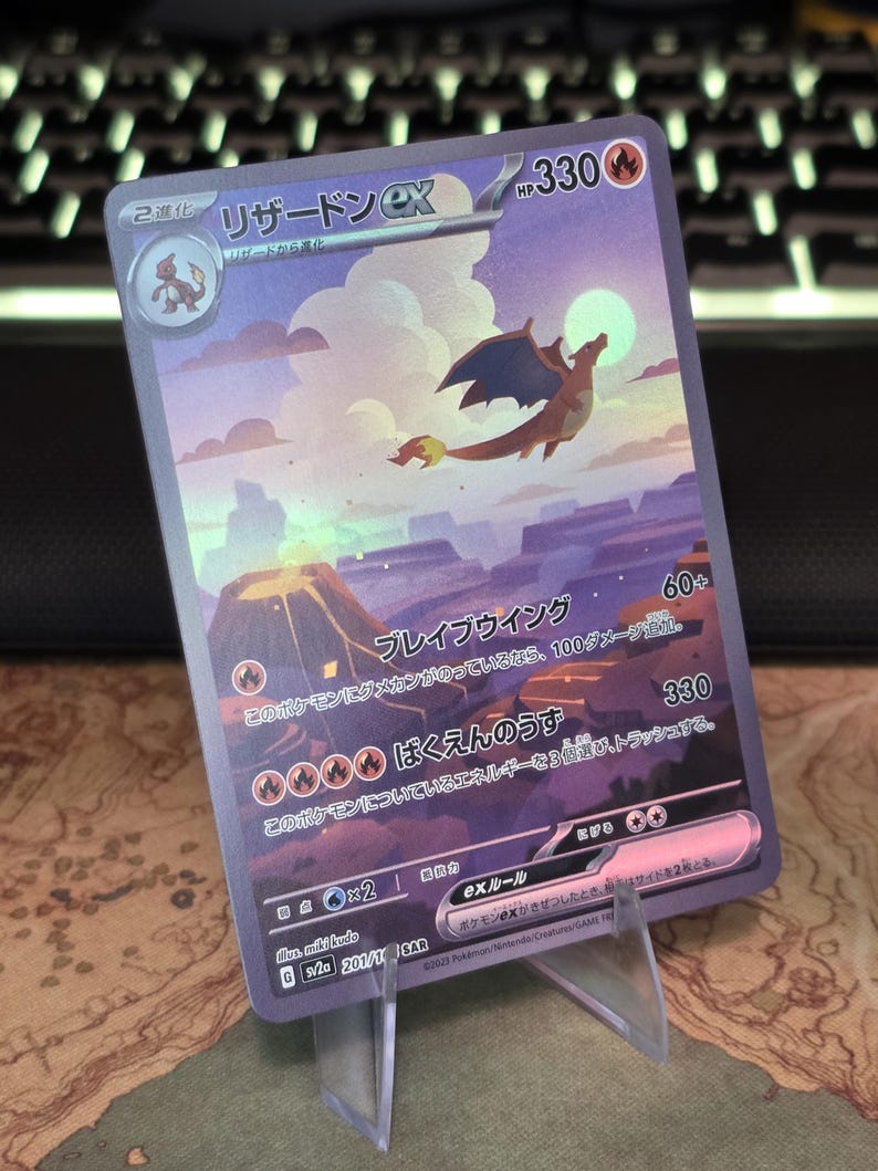 Pu&ograve; includere: Una carta collezionabile olografica Charizard ex con 330 HP, raffigurante un drago che vola sopra un paesaggio vulcanico al tramonto. La carta presenta testo in giapponese, simboli di energia fuoco e il codice del set sv2a 201/165 SAR.