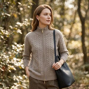 以下が含まれることがあります： ベージュのケーブルニットジップアップセーターを着た女性。ハイカラーとゴールドのジッパーが付いています。茶色のパンツと黒のレザートートバッグを持っています。背景はぼやけた森のシーンです。