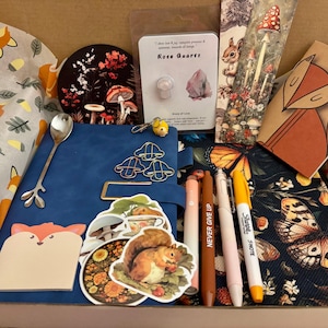 May include: A gift box containing stationery and decorative items. Includes a blue journal, pens, stickers, a bookmark, and a decorative spoon. The box has a woodland theme with fox and mushroom illustrations.