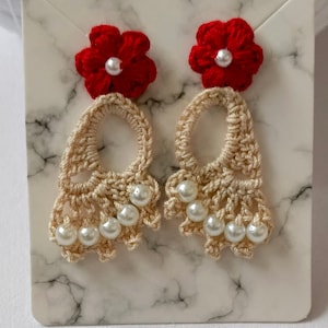 Puede incluir: Pendientes de ganchillo hechos a mano. Cada pendiente presenta una flor roja oscura con un centro de perla y un diseño beige de tejido abierto adornado con perlas de imitación. El texto en la tarjeta dice: "The petals are Dark Red."