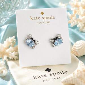 Peut inclure: Une paire de boucles d'oreilles clous en forme de crabe, dotées de pierres ovales bleu clair à facettes pour le corps et de petites pierres noires pour les yeux. Les boucles d'oreilles sont fixées sur un support blanc de la marque Kate Spade New York.