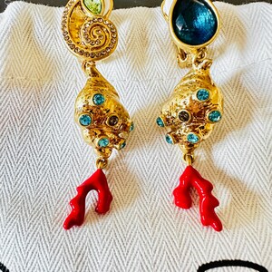 Pode incluir: Um par de brincos de pressão dourados com design em forma de concha. Um tem o topo em espiral com uma pedra verde-clara, o outro uma pedra azul-escura em forma de gota. Ambos possuem pequenos cristais azuis e um pendente de coral vermelho.