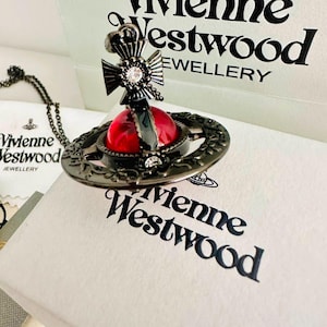 Può includere: Una collana con pendente orb Vivienne Westwood caratterizzata da una finitura metallica scura, una sfera centrale rossa traslucida e accenti in cristallo trasparente. Il design include una parte superiore a croce e una struttura ad anello dettagliata.