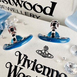 Pode incluir: Um par de brincos de tarracha prateados com um design de orbe azul e um emblema de cruz no topo, acentuados com pequenos cristais pretos. Os brincos estão expostos sobre uma caixa branca com o texto Vivienne Westwood Jewellery em preto.