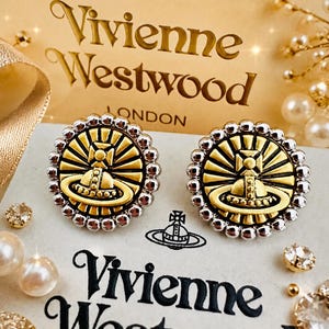 Peut inclure: Une paire de boucles d'oreilles rondes à clous présentant un emblème orbe doré sur un fond en rayons de soleil, entouré d'une bordure perlée argentée. Les boucles d'oreilles sont présentées sur une carte blanche avec le logo Vivienne Westwood.