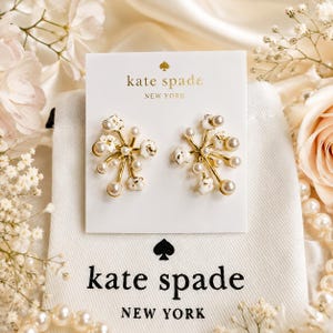 Peut inclure: Une paire de boucles d'oreilles à tige dorées avec des accents en forme de fleurs blanches et de petites perles artificielles rondes disposées en étoile. Elles sont présentées sur une carte blanche marquée Kate Spade New York.