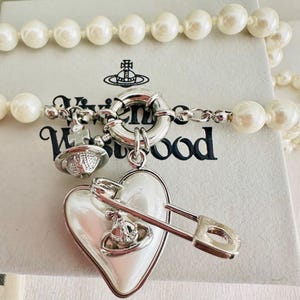 Peut inclure: Un collier de perles blanches avec un fermoir circulaire argenté. Un grand pendentif en forme de cœur blanc est attaché, orné d'une épingle à nourrice argentée et d'une petite breloque orbe argentée. Le logo Vivienne Westwood est sur la boîte.