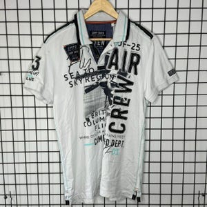 May include: A white short-sleeved polo shirt featuring a black collar with light blue trim. The front displays black graphic text including "AIR CREW," "JF-25," and "CAMP DAVID," along with a black and white image of an aeroplane propeller.