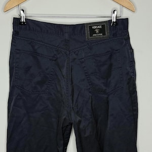 May include: A pair of dark navy blue trousers featuring two back pockets and a black rectangular patch on the waistband. The patch displays the text VERSACE JEANS COUTURE along with a small graphic of a head centered above the brand name.